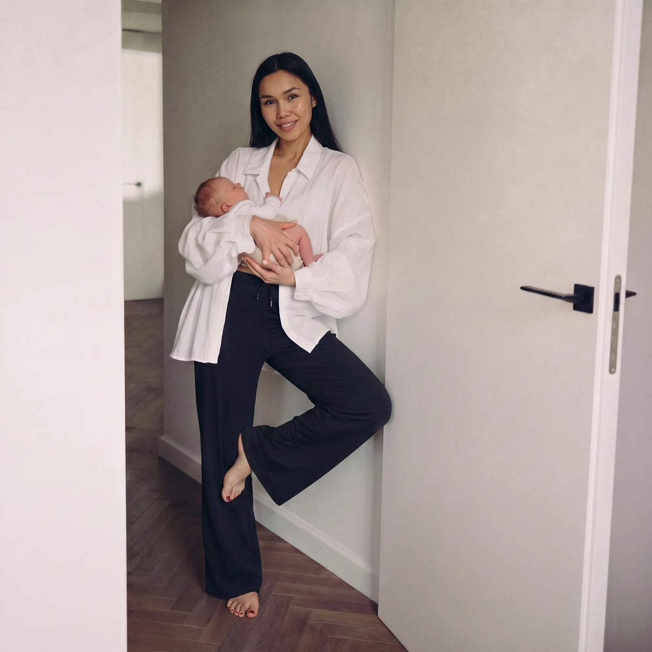 A woman in a white button-down shirt and black wide-leg pants holds a sleeping newborn baby in a doorway of a home interior.