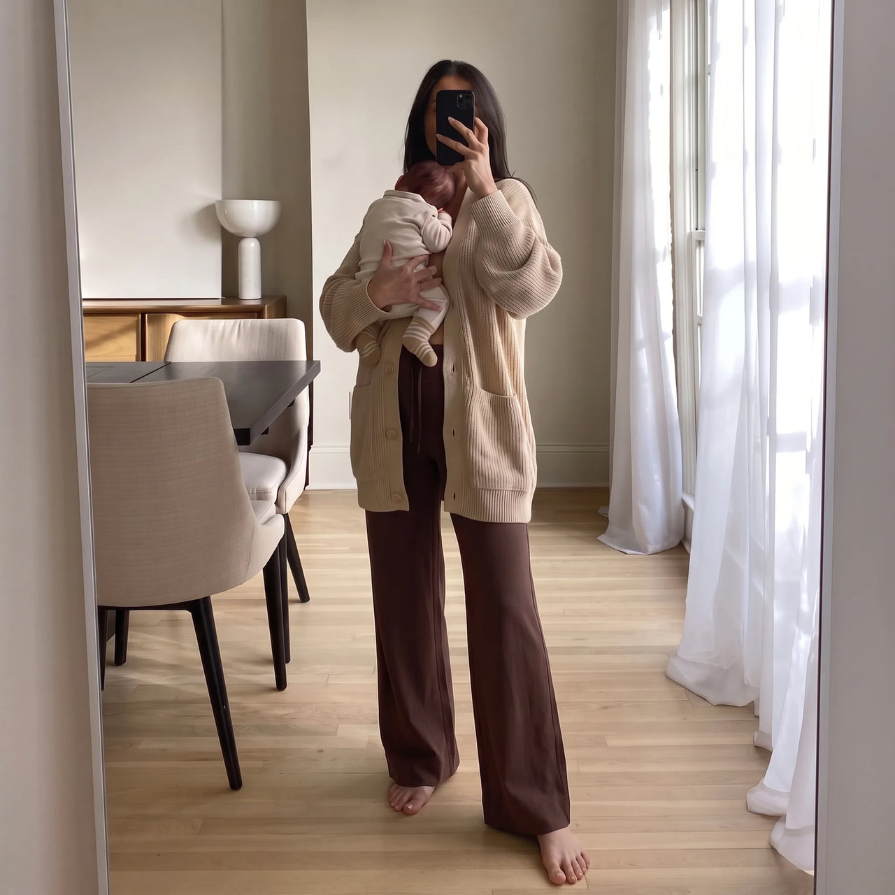 A woman takes a mirror selfie in a bright room while holding a baby. She is wearing a beige knit cardigan, brown wide-leg pants, and a cream baby outfit.