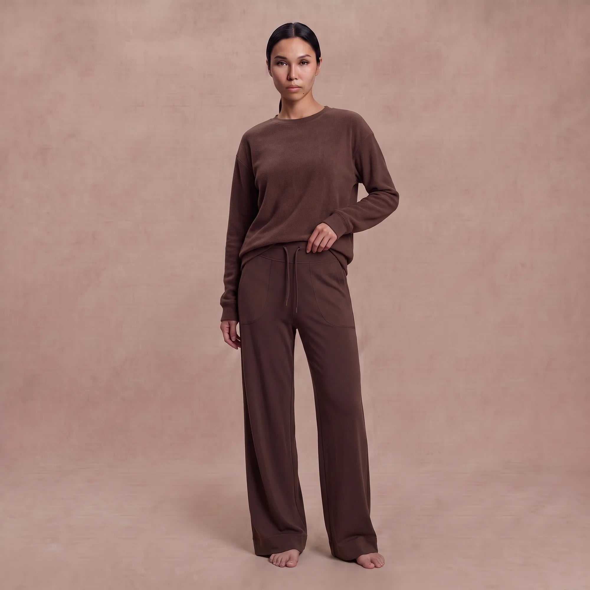 A woman stands barefoot against a dusty pink studio backdrop, wearing a matching brown lounge set consisting of a long-sleeve crewneck top and wide-leg drawstring pants.
