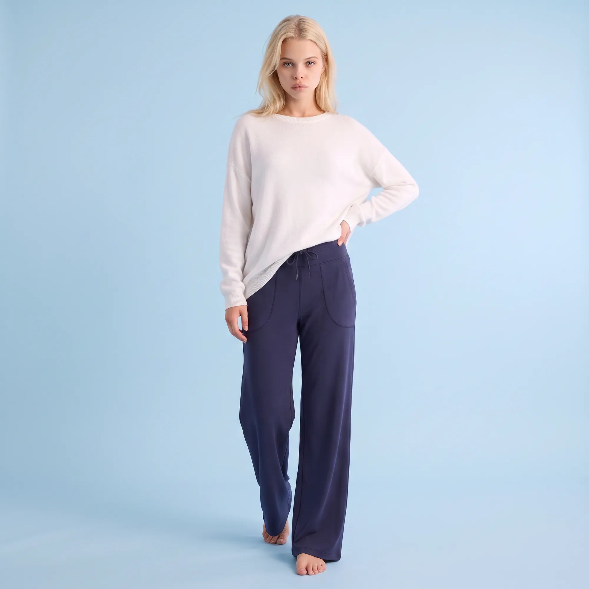 A woman stands barefoot against a light blue studio background, wearing a loose off-white long-sleeve top and dark navy wide-leg pants with front patch pockets.