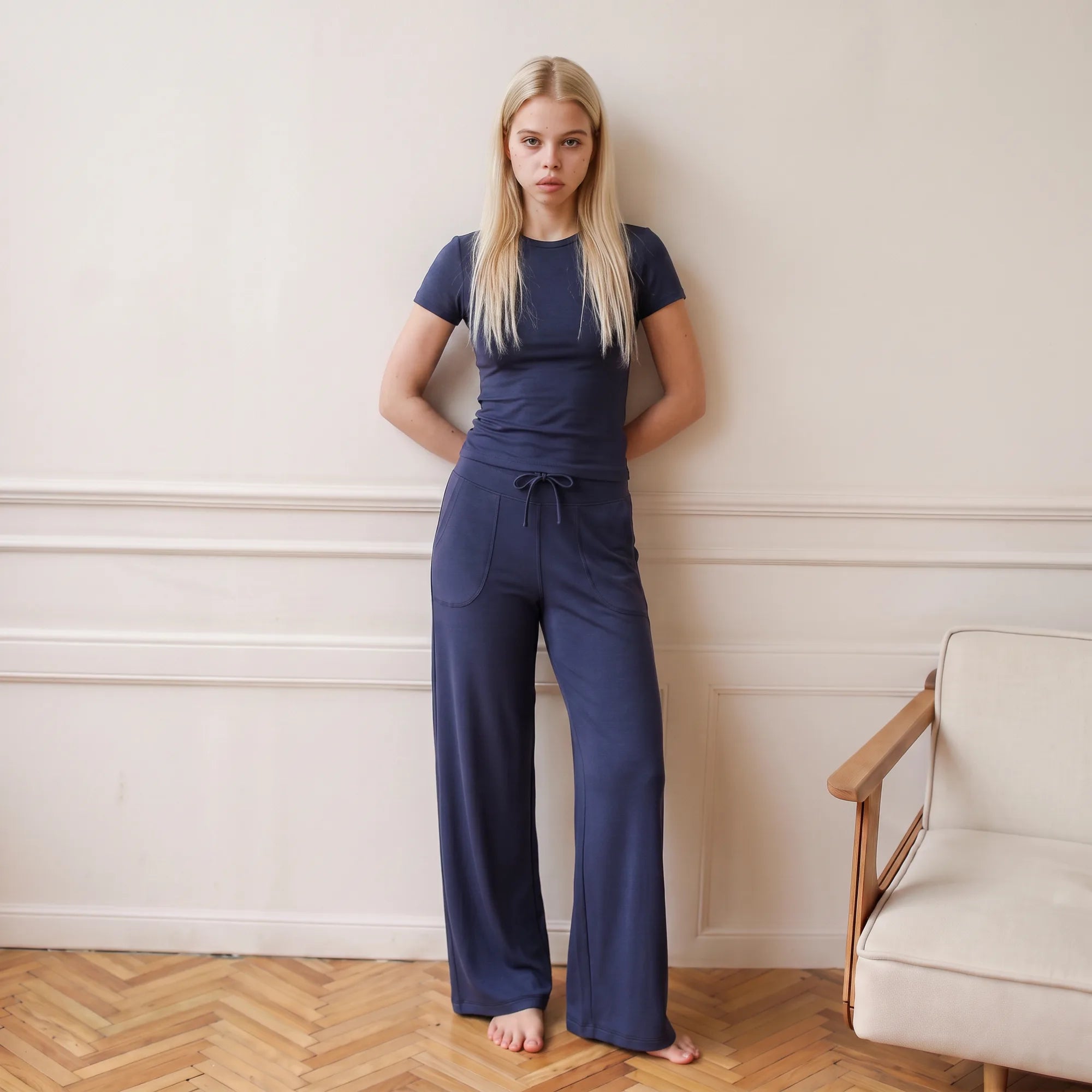 A woman stands barefoot against a cream-colored wall with molding, wearing a navy blue short-sleeve t-shirt and matching wide-leg pants. A light-colored armchair is partially visible on the right over a herringbone wood floor.